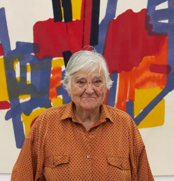 Etel Adnan standing in front of one of her abstract paintings, wearing a rust-colored polka-dot shirt. The vibrant artwork behind her features bold red, blue, and yellow forms, reflecting her signature use of color and geometric abstraction.