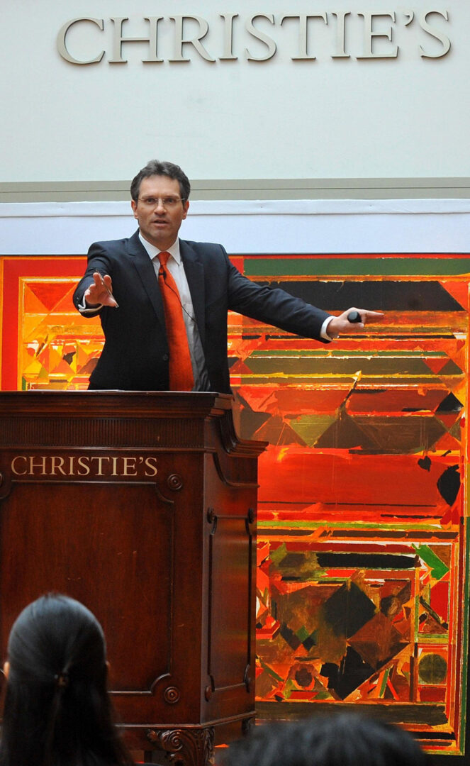 An auctioneer at Christie’s conducting a live auction, with a vibrant artwork displayed in the background, reflecting the high-stakes art market where Sayed Haider Raza’s works have been sold for record prices.