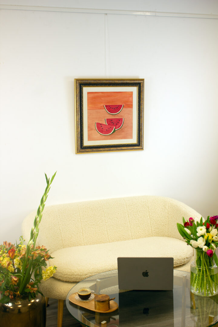 "Sandías, 1972" by Rufino Tamayo displayed on a white wall in an interior setting with a modern sofa, laptop, and flowers.