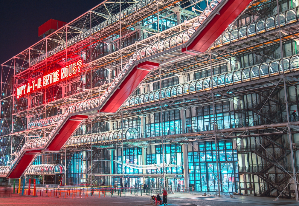 The Centre Pompidou in Paris at night, featuring its modern architecture with colorful pipes and a large illuminated sign displaying the text "Qu'y a-t-il entre nous?" ("What is there between us?")