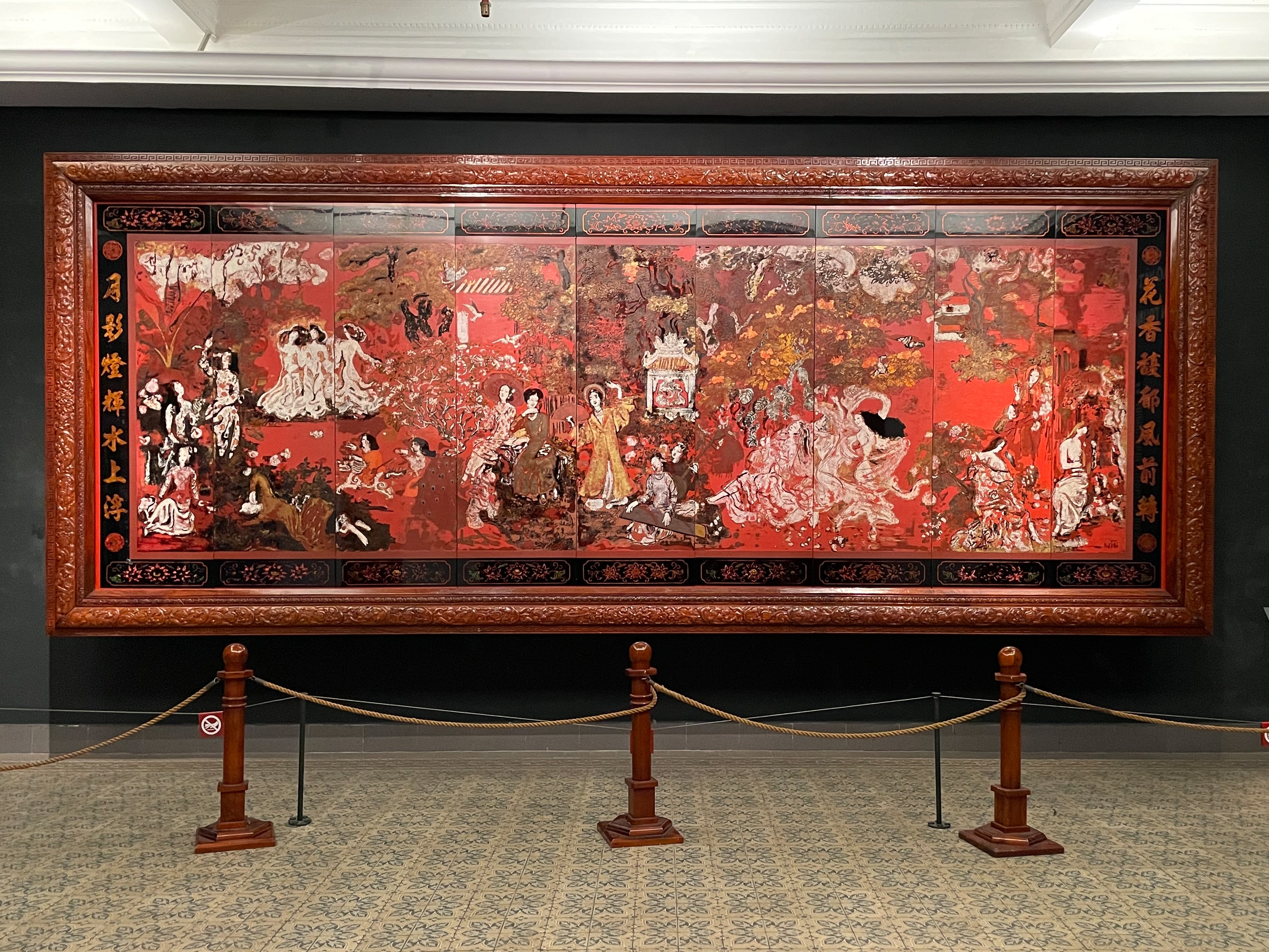 A grand lacquer painting by Nguyen Gia Tri, a master of 20th century asian art, displayed in an ornate wooden frame at a museum. This artwork exemplifies the fusion of traditional Vietnamese lacquer techniques with modernist expression.