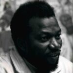 Close-up black-and-white portrait of Ben Enwonwu smiling, wearing a light-colored coat, with a blurred background of his studio or paintings.