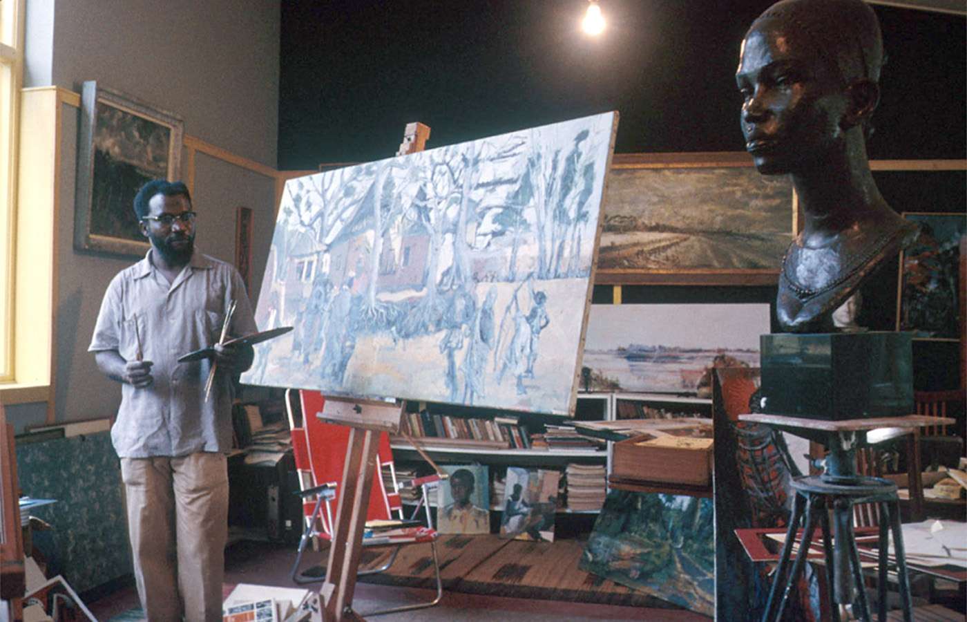 Ben Enwonwu painting in his studio, surrounded by works in progress and a large bronze bust, reflecting his multifaceted artistic practice. | Aurora & Athena Auction House Ben Enwonwu painting in his studio, surrounded by works in progress and a large bronze bust, reflecting his multifaceted artistic practice.