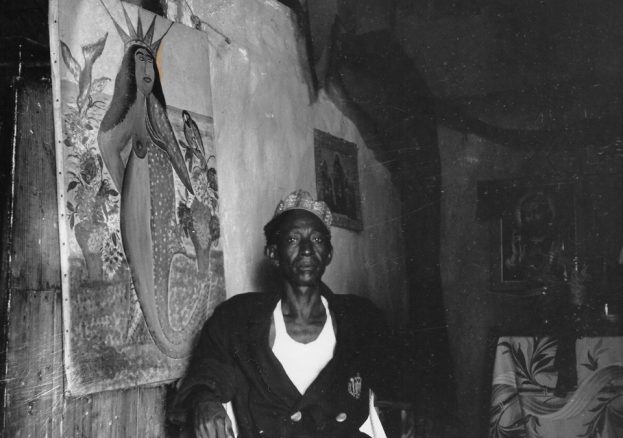 Photograph of Haitian artist Hector Hyppolite seated beside his painting of La Sirène, a Vodou mermaid goddess – a rare glimpse into the legendary painter’s spiritual studio.