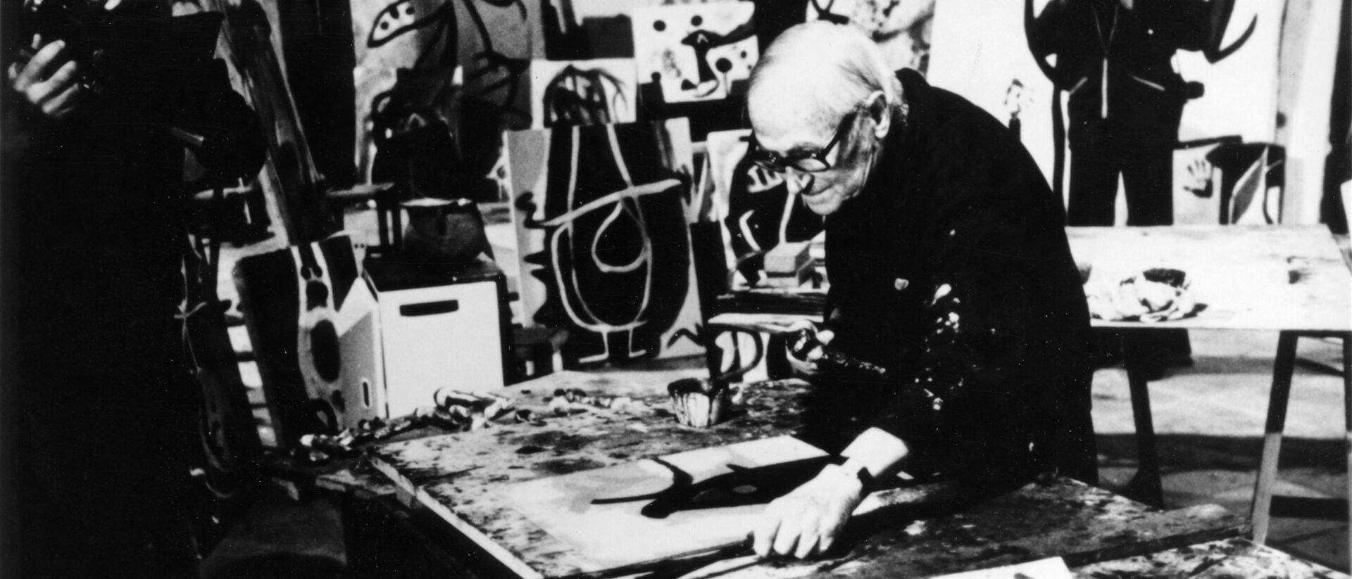Vintage black-and-white photograph of Spanish Surrealist master Joan Miró working on a painting in his studio. Discover authentic Miró original artworks for sale at Aurora & Athena gallery